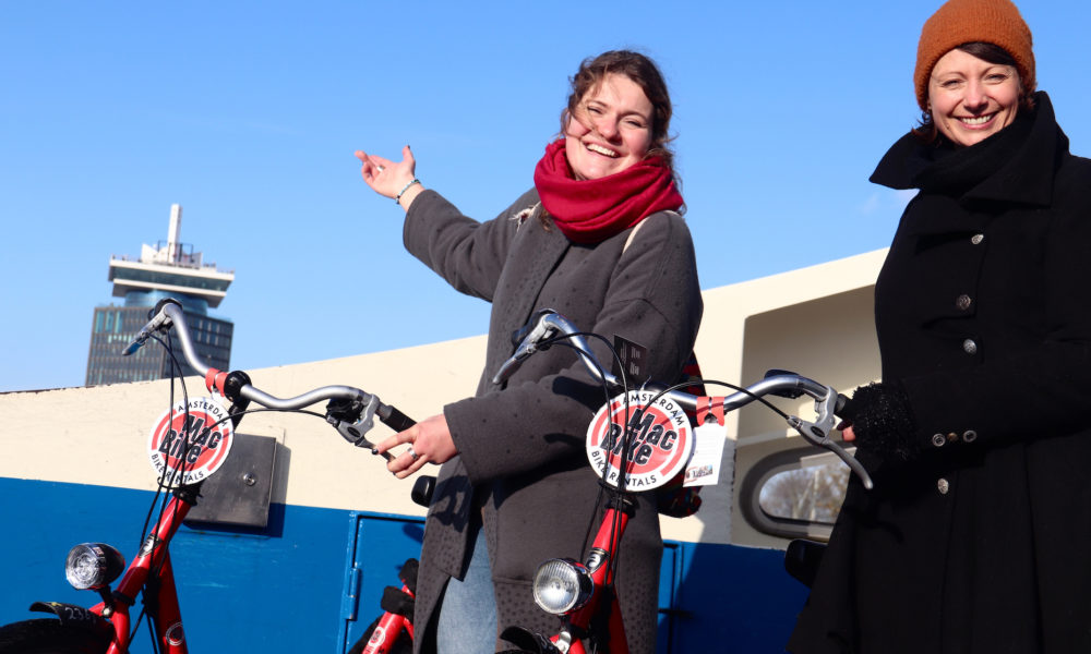 Two women smiling on e-bikes in Amsterdam. They are enjoying a sunny day.