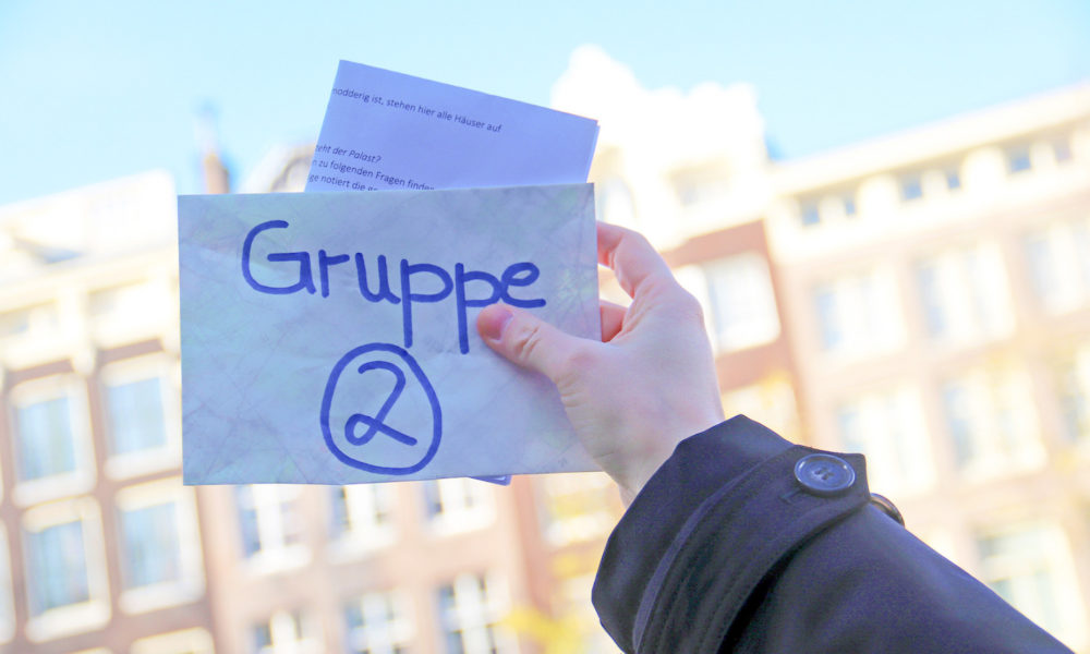 Hand holding a card with Gruppe 2 written on it. A building in Amsterdam is in the background.