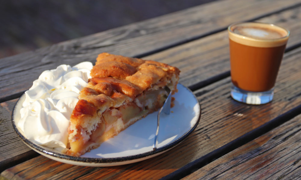 A slice of apple pie with whipped cream on a plate and a cup of coffee on a wooden table.