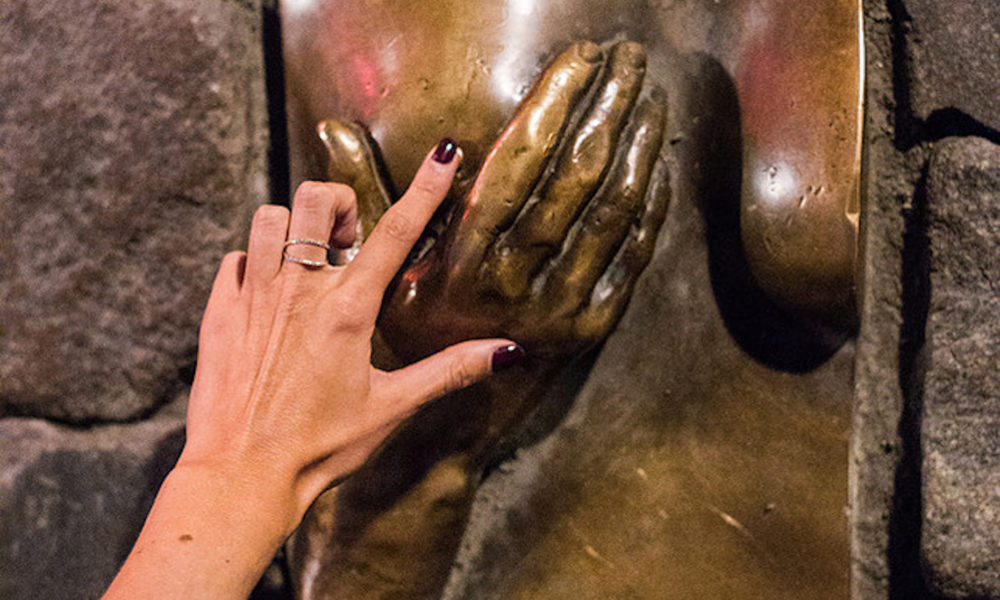 A woman's hand touching a bronze statue. The statue is likely a famous Amsterdam landmark.