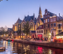 A serene Amsterdam canal at dusk with boats and buildings. People enjoy the evening.