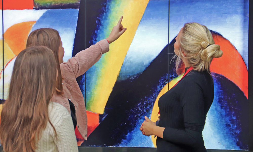 Women looking at a vibrant abstract artwork. The painting features bold colours and geometric shapes.