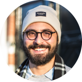 Portrait of a man with a beard wearing a white beanie and glasses. He appears to be enjoying a city trip.