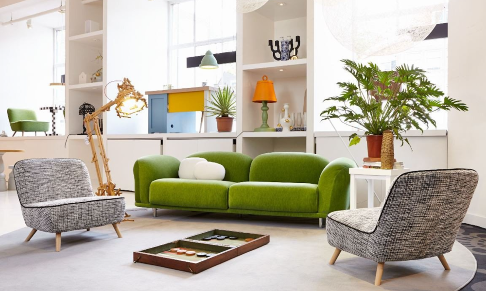 A modern living room with a green sofa and two patterned chairs. The room is bright and airy with large windows.