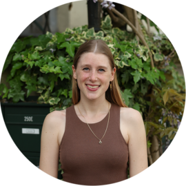 A smiling woman with long brown hair, wearing a brown tank top and a necklace. She is standing in front of green plants.