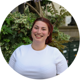 A smiling woman with red hair standing in front of lush greenery. She is wearing a white shirt.