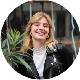 A young woman with blonde hair smiling. She is wearing a white shirt and a black leather jacket.