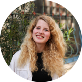 A young woman with long blonde hair smiling. She is standing in front of some plants.