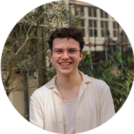 A smiling young man standing outside with plants behind him. He is wearing a white shirt.