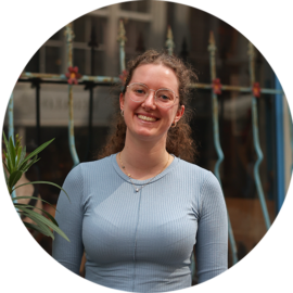A young woman with brown hair wearing a blue top smiling at the camera. She appears to be outdoors.